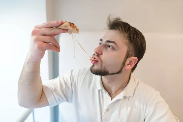A handsome man eats a hot piece of pizza. The man eats a cheese that stretches out with a slice of pizza. — Stock Photo, Image