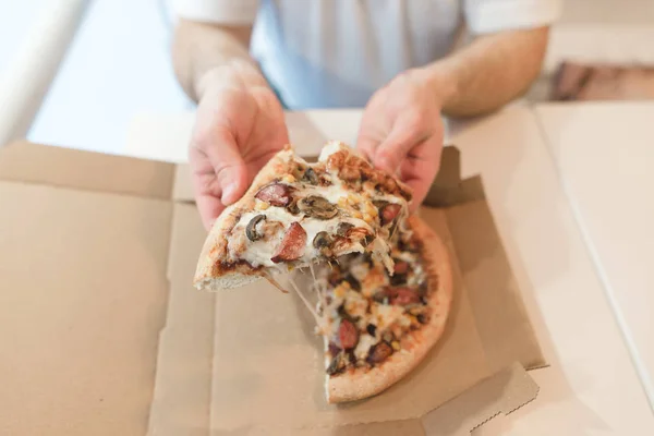 Trozos de pizza apetitosa en las manos de los hombres. Un hombre come una pizza caliente de una caja de papel . — Foto de Stock