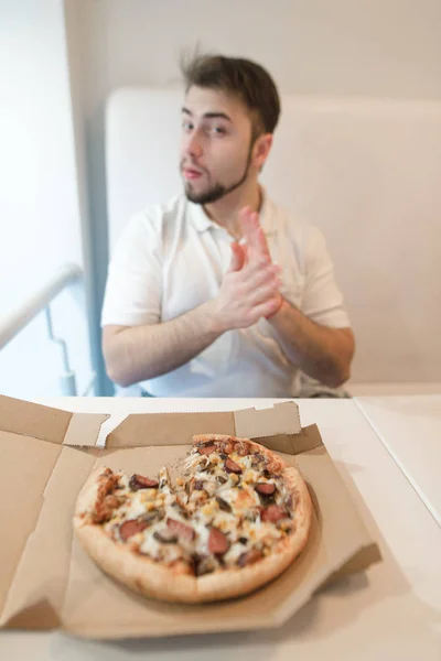 Mann sitzt an einem Tisch in der Nähe der Pizzakiste und blickt in die Kamera. Schwerpunkt Pizza. Pizza auf dem Hintergrund eines Mannes. — Stockfoto