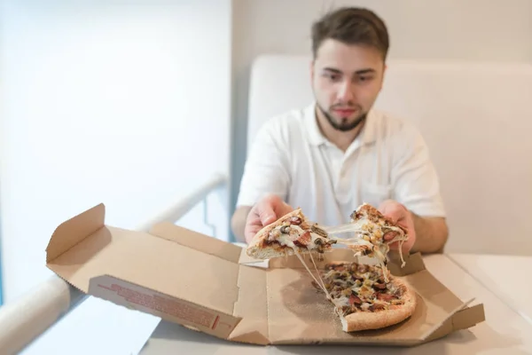 Чоловік сидить за столом біля коробки і тримає в руках кілька шматків піци. Чоловік їсть гарячу піцу . — стокове фото