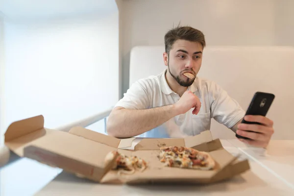 Um homem bonito e engraçado come uma pizza da caixa e pega seu telefone. Selfies com pizza . — Fotografia de Stock