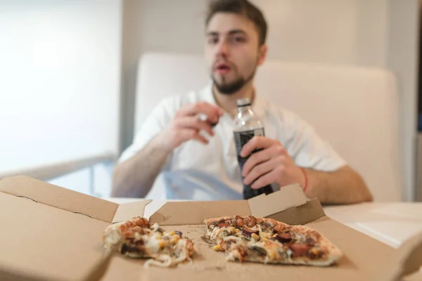 Ein verschwommener Mann mit einem Getränk in der Hand gegen eine Pizzakiste. Der Mann isst die Fasnet und trinkt die Cola. Der Mann isst eine Pizza. — Stockfoto