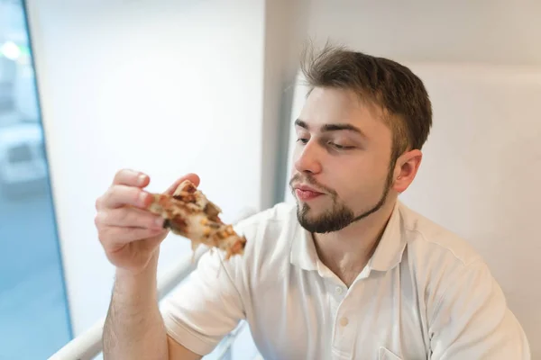 Un hombre guapo mira de cerca un pedazo de pizza en sus manos. El estudiante come pizza para el almuerzo. Un pedazo de deliciosa comida rápida. Concéntrate en el hombre . —  Fotos de Stock