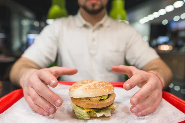 Um homem come um hambúrguer grande que está em pé na bandeja. Mãos no ar — Fotografia de Stock