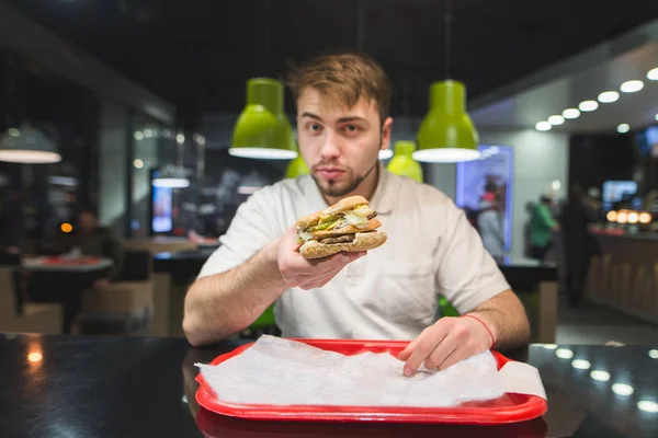Een knappe man houdt van een hamburger in zijn handen en stuurt hem naar de — Stockfoto