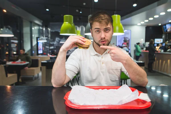 Een man zit in een bistro en toont een patch op een hamburger in zijn handen. Een man met een chizburger kijkt naar de camera. Fast-food concept. — Stockfoto