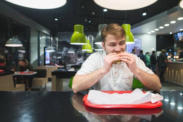 Голодный человек ест фаст-фуд в ресторане. Человек жадно ест бургер за столом у подноса. Концепция быстрого питания . — стоковое фото