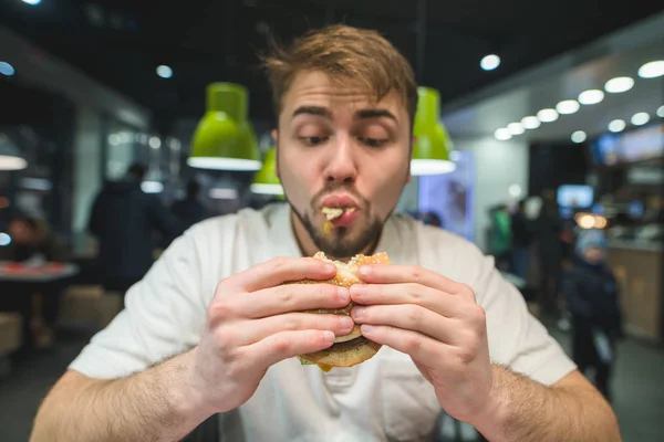 Un hombre hambriento con barba come codiciosamente una hamburguesa. Un hombre gracioso mirando un sándwich. Concéntrate en la hamburguesa. Concepto de comida rápida . — Foto de Stock