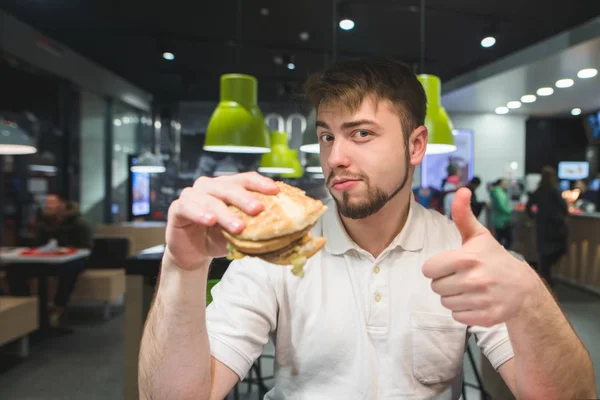 Een man met een hamburger in zijn handen zit in een fast-food restaurant en toont een thumbs up. Een man houdt van een hamburger. — Stockfoto
