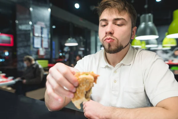 Um homem faminto senta-se em um restaurante de fast-food e olha para o hambúrguer em sua mão. Conceito de fast-food . — Fotografia de Stock