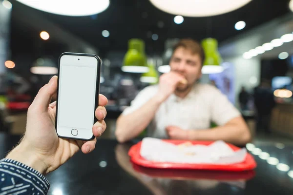 Ponsel dengan layar putih di belakang seorang pria yang makan makanan cepat saji di restoran. Makanan cepat saji untuk makan siang . — Stok Foto