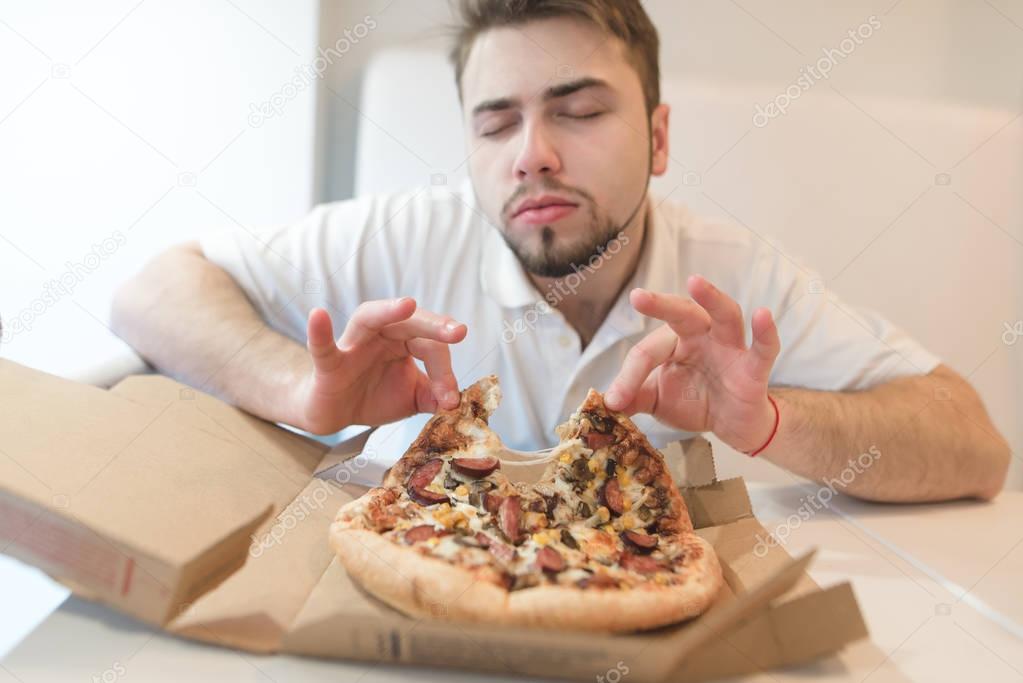 A nice man gets pleasure from the aroma of pizza. A man with his eyes closed takes a piece of pizza from a cardboard box