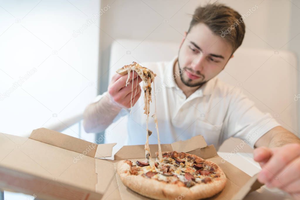 A handsome man holds a slender of appetizing pizza in his hands and stares at her with a hungry look. The man eats a hot pizza from a cardboard box.