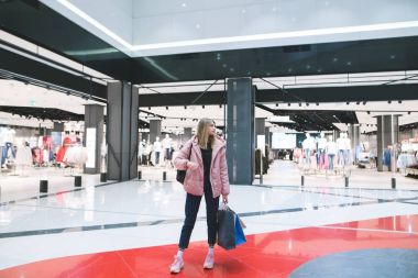 Onun elinde alışveriş torbaları ile şık bir sarışın kız bir giyim mağazası çerçevede alışveriş merkezinde duruyor.