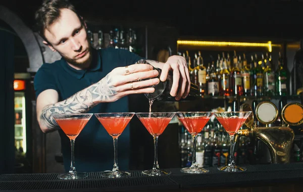 The barman from the tattoo makes beautiful alcoholic cocktails at the bar counter. The barman creates red women's cocktails at a nightclub bar — Stock Photo, Image
