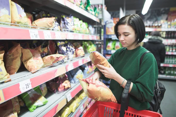 Una joven compra bocadillos en un supermercado. La chica elige fichas en un supermercado y las pone en un carrito de compras rojo. Compras en un supermercado . — Foto de Stock