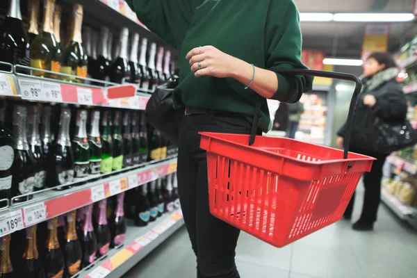Μια γυναίκα με μια κόκκινη τσάντα ψώνια επιλέγει αλκοόλ σε ένα σούπερ μάρκετ. Επιλογή των προϊόντων στο σούπερ μάρκετ. Η επιλογή του κρασιού στο κατάστημα. — Φωτογραφία Αρχείου
