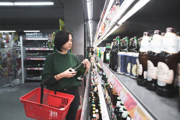 Een meisje met een rode mand kiest alcohol in een supermarkt. Winkelen voor bier in de winkel. Het meisje is winkelen in de supermarkt. — Stockfoto