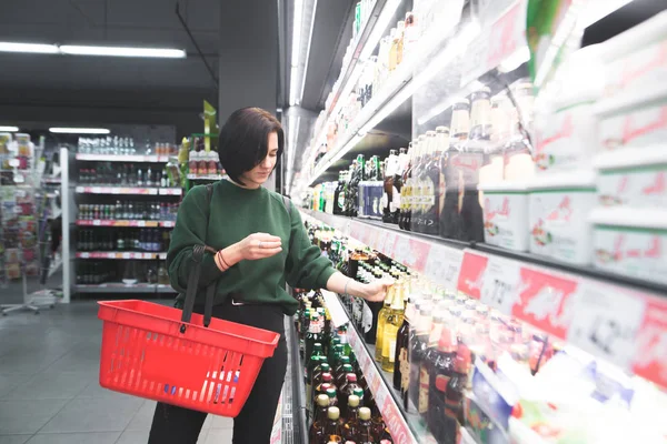 Uma rapariga sorridente tira cerveja da prateleira do supermercado. Uma rapariga bonita compra uma cerveja num supermercado. O comprador compra álcool na loja . — Fotografia de Stock