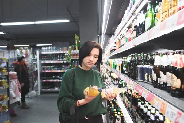 Pigen ser på ølflasker i sine hænder. En pige vælger en alkoholisk drik i et supermarked. Valget af alkohol i butikken - Stock-foto