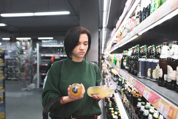 Portret dziewczynki, która wybiera piwa w supermarkecie. Dziewczyna patrzy alkoholu w jego ręce. Zakupy w supermarkecie. — Zdjęcie stockowe