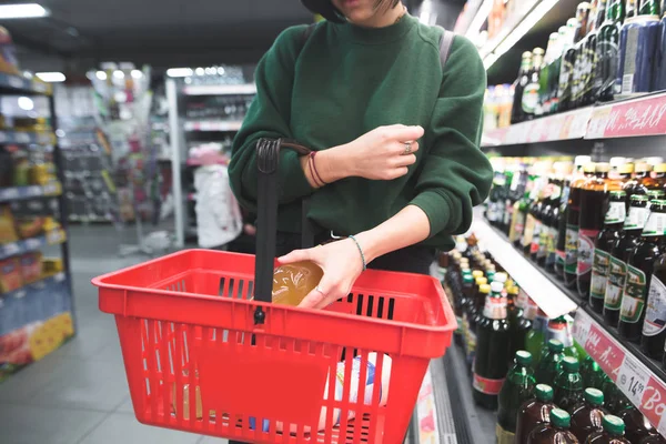 Una mujer pone una botella de alcohol en un carrito de compras rojo. La chica pone una compra en la cesta. Compras en un supermercado . —  Fotos de Stock