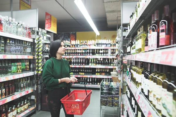 Krásná mladá dívka vybere víno v supermarketu. Kupující kupuje alkohol v supermarketu. Podívejte se na police supermarketu. — Stock fotografie