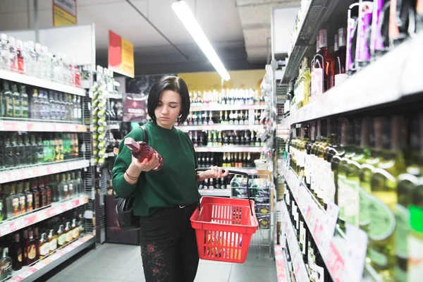 Portret dziewczynki, która wybiera alkoholu w supermarkecie. Jej mąż patrzy na butelkę wina w czasie zakupów. Zakupy w supermarkecie — Zdjęcie stockowe