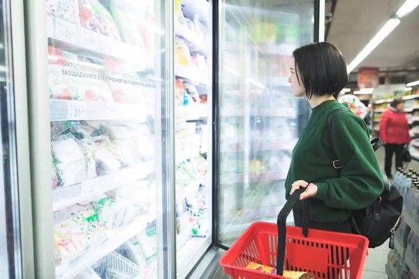 Seorang gadis dengan keranjang belanja membuka lemari es untuk makanan beku di supermarket. Gadis itu berbelanja di supermarket . — Stok Foto