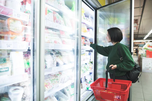 Uma rapariga com uma cesta tira alimentos congelados do frigorífico do supermercado. A menina escolhe mercadorias na loja. Compras no supermercado . — Fotografia de Stock