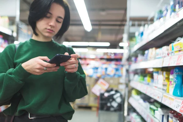 Dziewczyna używa smartphone przy wyborze towaru w supermarkecie. Zakupy w supermarkecie. — Zdjęcie stockowe