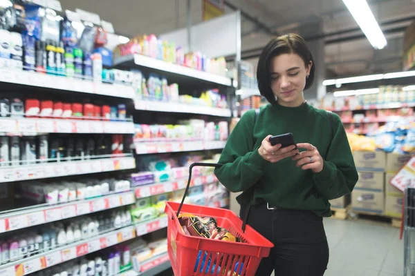 Pozytywna dziewczyna wykorzystuje telefon w supermarkecie. Dziewczyna zakupy w supermarkecie z telefonem w ręce. Skupić się na Twój telefon — Zdjęcie stockowe