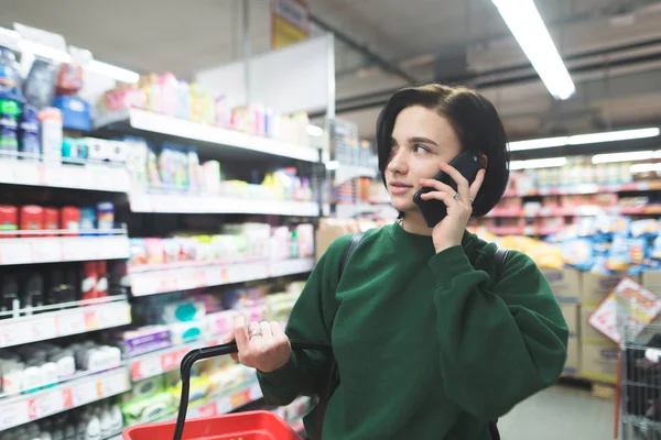 Frumoasă fată tânără vorbind la telefon la supermarket și arătând lateral. Fata face cumpărături la supermarket . — Fotografie, imagine de stoc