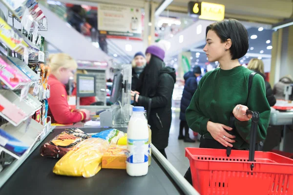 Krásná dívka v supermarketu stojí a čeká na frontu. Částka platí pro nákupy na pokladně v supermarketu. — Stock fotografie