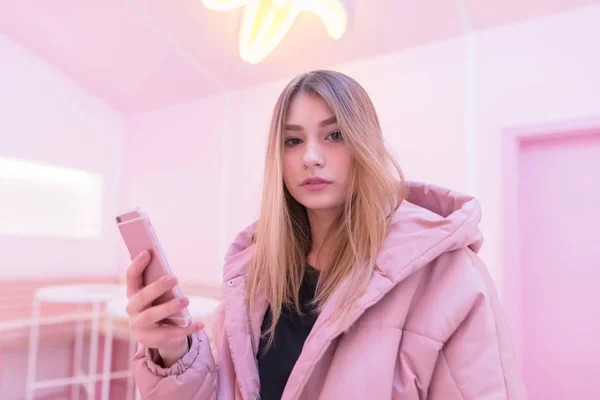 The girl in pink clothes uses a pink smartphone in a room with pink interior. Pink life A look at the camera.