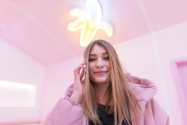 Uma garota positiva em roupas rosa falando no telefone em uma sala rosa e olhando para a câmera. Vida rosa — Fotografia de Stock