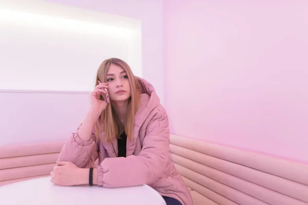Mooi blond meisje zit aan de tafel op de roze zitbank en spreekt telefonisch in een kamer met roze interieur. Schattig meisje kijkt naar de kant — Stockfoto