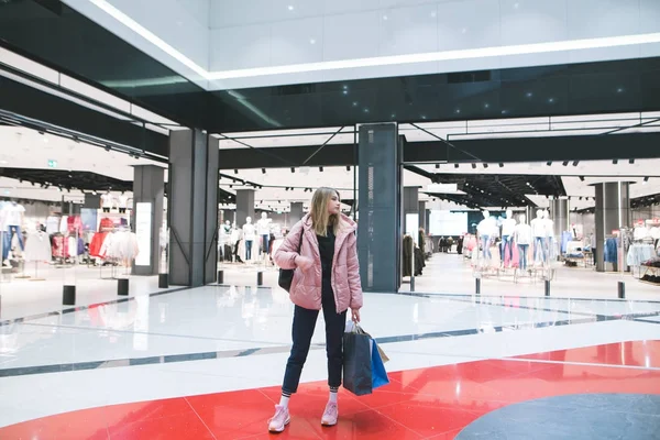 En snygg blond tjej med shoppingkassar i händerna står i gallerian mot bakgrund av en klädaffär. — Stockfoto