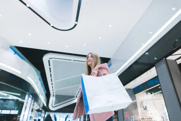 Une blonde douce avec des sacs à provisions dans les mains d'un centre commercial regardant dans la caméra. Shopping dans un centre commercial moderne . — Photo