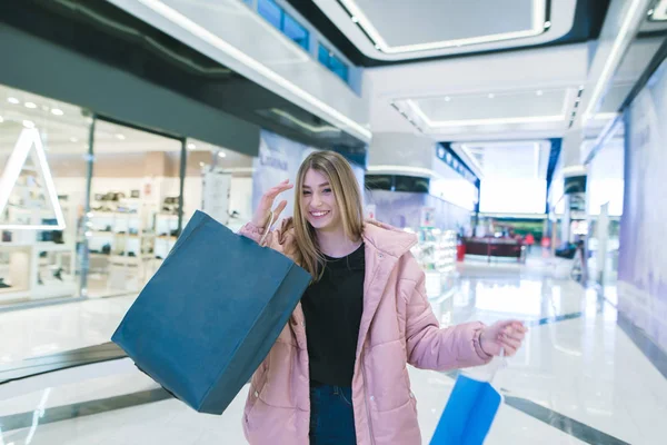 En smilende pige med shopping i hænderne på et ægte indkøbscenter. Indkøbskoncept . - Stock-foto