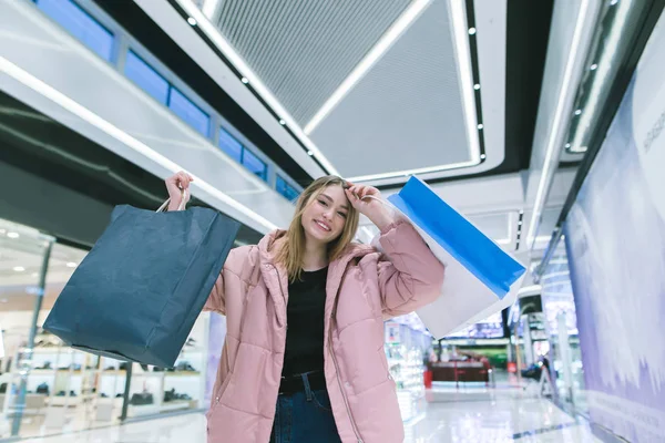En leende blondin i en vacker shoppinggalleria uppreser shoppingkassar. Shopping koncept. — Stockfoto