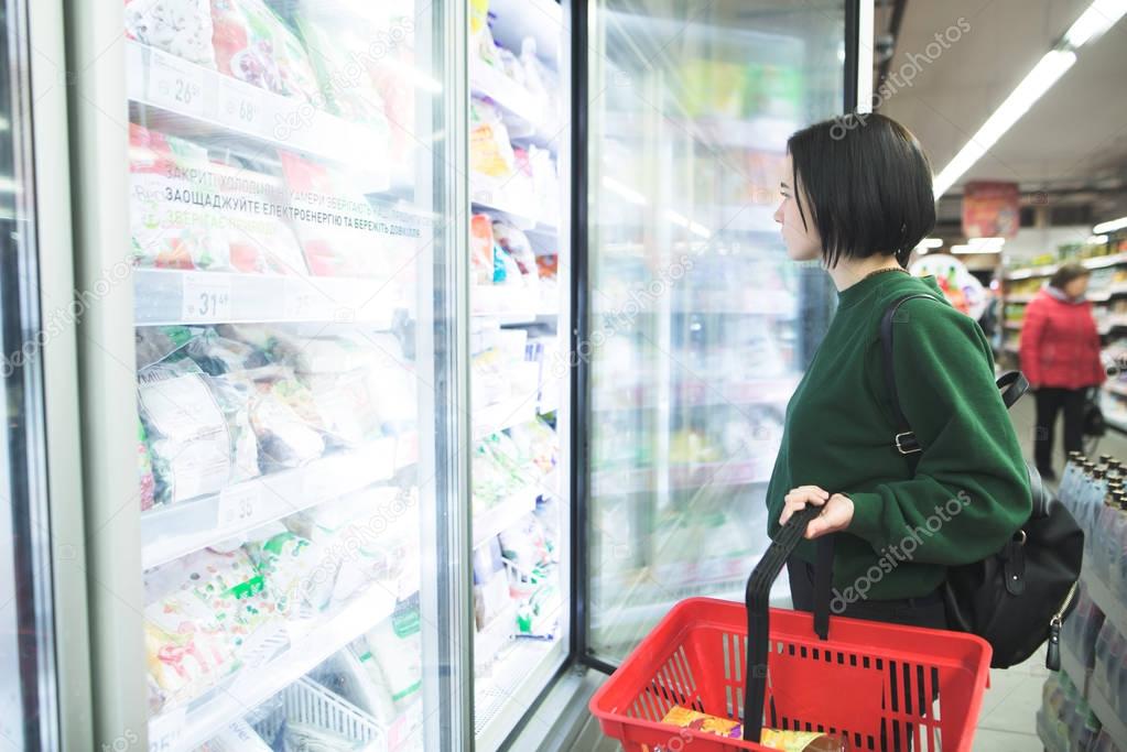 A girl with a shopping cart opens a fridge for frozen foods in the supermarket. The girl is shopping at the supermarket.