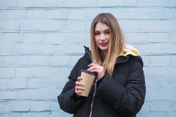 Menina elegante positivo com um copo de bebida quente em pé na rua perto da parede azul e olhando para a câmera — Fotografia de Stock