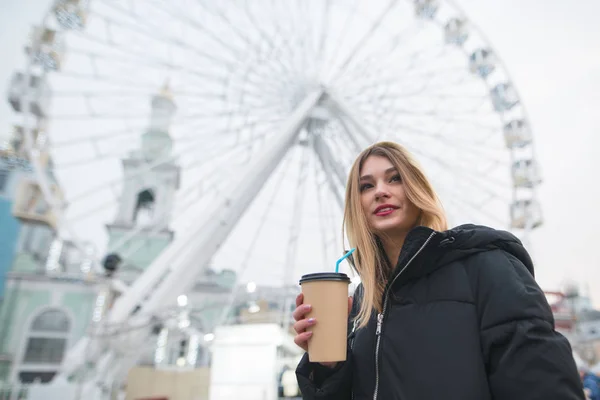 Porträt eines schönen positiven Mädchens mit einer Tasse Kaffee in der Hand vor dem Hintergrund eines Riesenrads. — Stockfoto