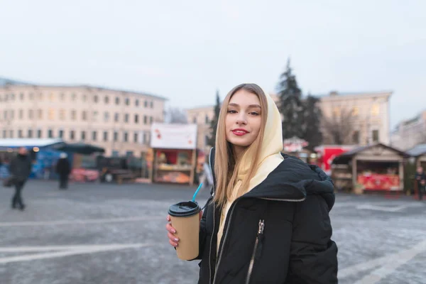 Uma garota positiva com um copo de café nas mãos fica no fundo da feira, sorri e olha para a câmera . — Fotografia de Stock