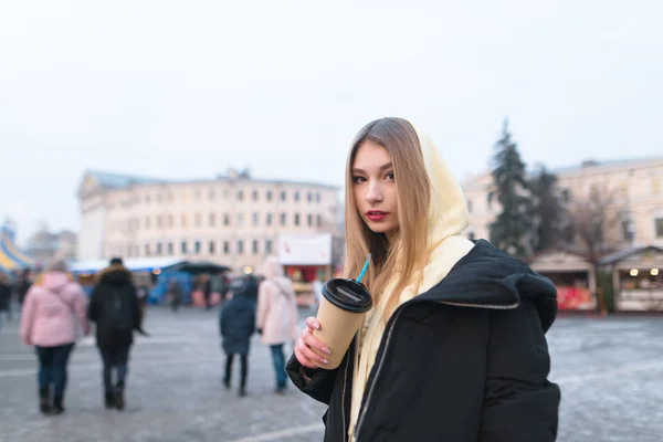 美しい少女は、彼女の手の通り市の冬の一杯のコーヒーでバック グラウンドで立っているし、カメラを見ています。女の子は温かい飲み物と暖かい — ストック写真