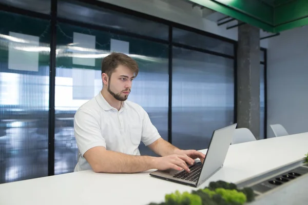 Un uomo con la barba lavora in un portatile in ufficio. Un giovane uomo gode di un computer in un ufficio moderno . — Foto Stock