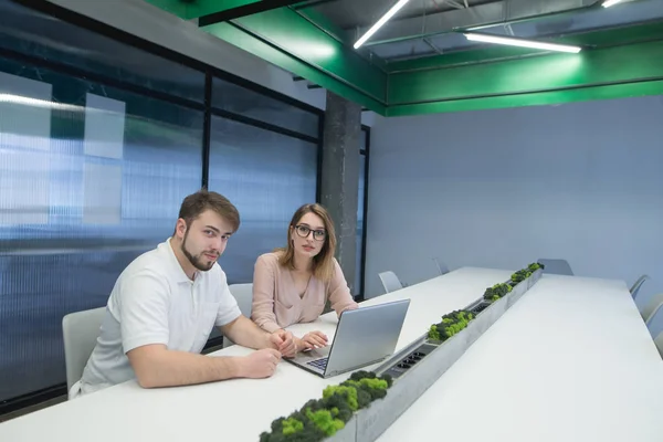 Un homme et une femme sont assis au bureau près du bureau au tableau noir et regardent la caméra. Les jeunes employés de bureau mettent la caméra dans l'espace de travail moderne . — Photo