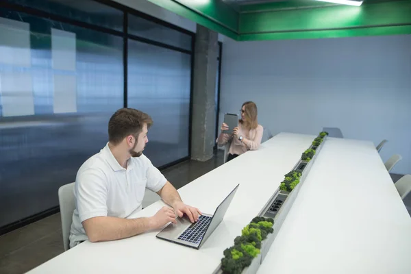 Un uomo con la barba lavora su un portatile nell'area di lavoro, e una donna fotografa su un piatto. Seduto in ufficio. Uomo con una donna sul posto di lavoro . — Foto Stock