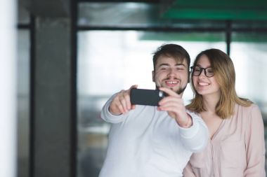 Gülümseyen adam ve kız selfie kapalı olun. Selfie akıllı telefon yapmak yazıhane amele güzel çifti. Mutlu çalışanlar bir smartphone cep telefonu ile ofiste fotoğraflandı.